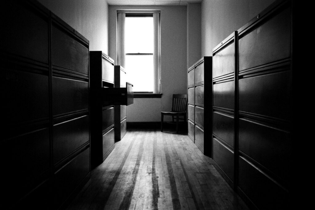 Couloir de bureau en noir et blanc bordé de classeurs métalliques, une chaise vide face à la fenêtre — audit social